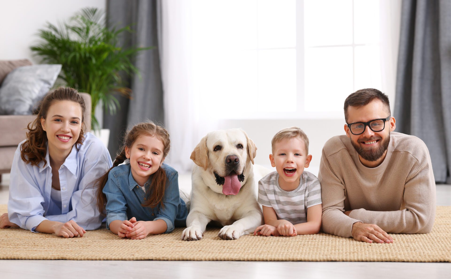 Happy idyllic family with kids and dog resting at home Smiling mother and father with little kids and adorable purebred Labrador retriever dog lying on floor and looking at camera while spending time together at home